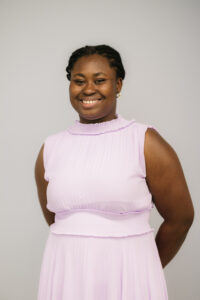 Mercy, a young Black woman with braids pulled back, smiles at the camera. She is wearing a lavender-pink dress and small gold hoop earrings.  