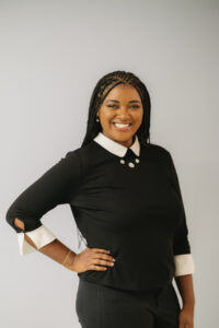 Image description: Brynn, a Black woman in her 20s with long, curly hair styled in box braids, stands in front of a grey background, smiling brightly at the camera with one hand on her hip. She is wearing a black top with a white collar and sleeves, with delicate pearl earrings, gold bracelet, and decorative necklace.
