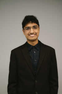 Rohit, a South-Asian Indian man with short curly black hair and eyeglasses without frames, smiles at the camera indoors against a grey background. He wears a black blazer with a black button-down shirt. 