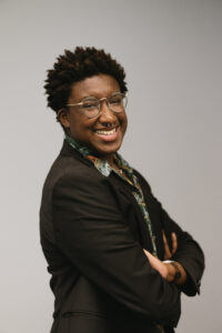 Malcolm, a Black person with dark hair and wire-rimmed glasses is smiling and crossing his arms in an ornate green button-down and black suit jacket.