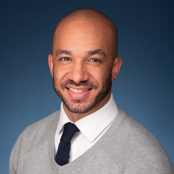 Professional headshot of a smiling light-skin Black man with a bald head and trimmed beard, wearing a light gray sweater over a white collared shirt and navy blue tie, set against a dark blue background.