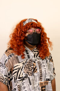 A headshot of Marrow, a Black person with curly red hair posing against a neutral backdrop. They are wearing a black face mask, oversized round glasses with rose-tinted lenses, and a black-and-white patterned headband. Their button-up shirt is patterned with newspaper clippings and animal prints, blending text with zebra and leopard motifs.