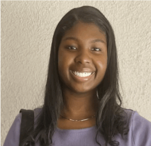 ID: Makayla, a Black mixed woman with medium length black hair. She wears a silver necklace with a purple shirt and black camera bag strap.